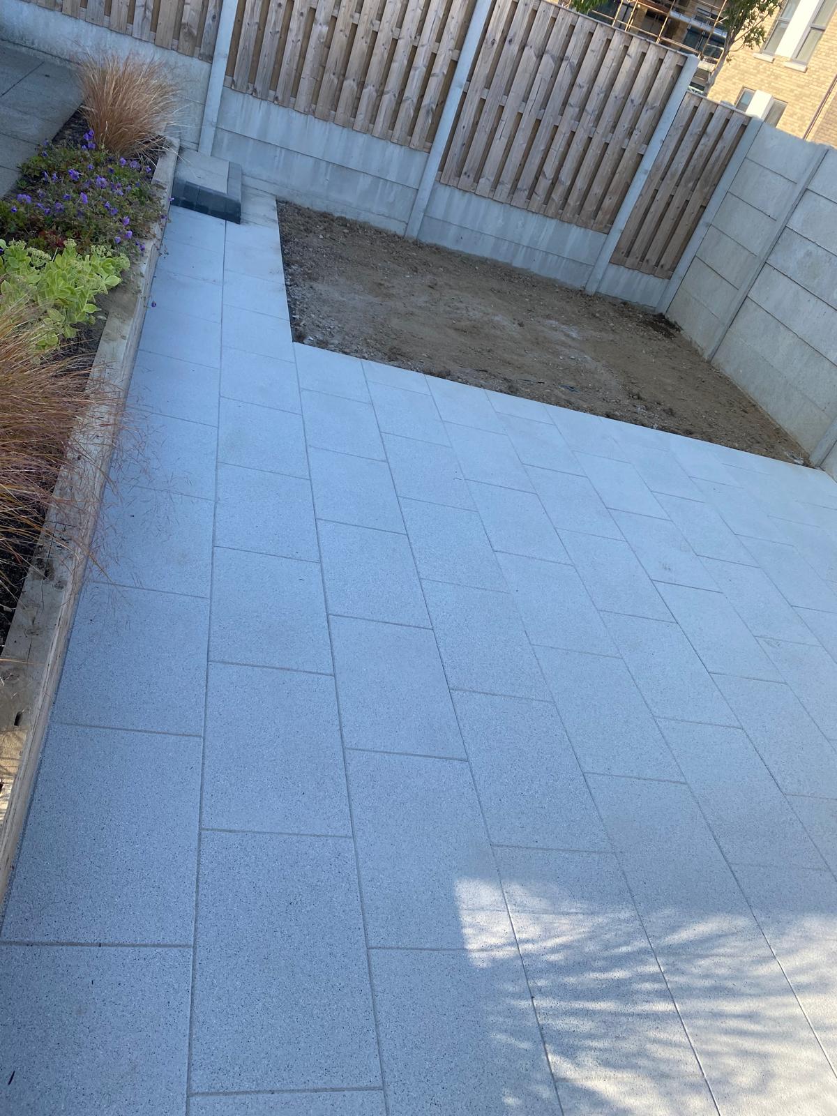 Pale granite slab patio laid in a running bond beside a timber sleeper raised bed of grasses and flowering planting, next to a prepared soil area and fencing