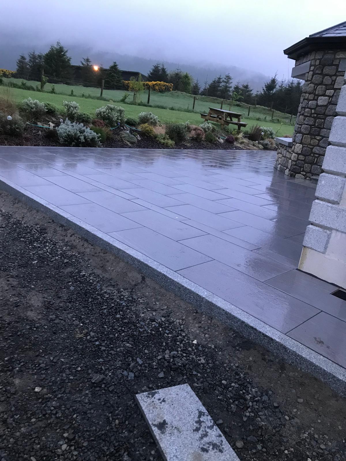 Dark large format paved patio with a light granite edge course beside a stone clad pillar, overlooking planting and hillside fields
