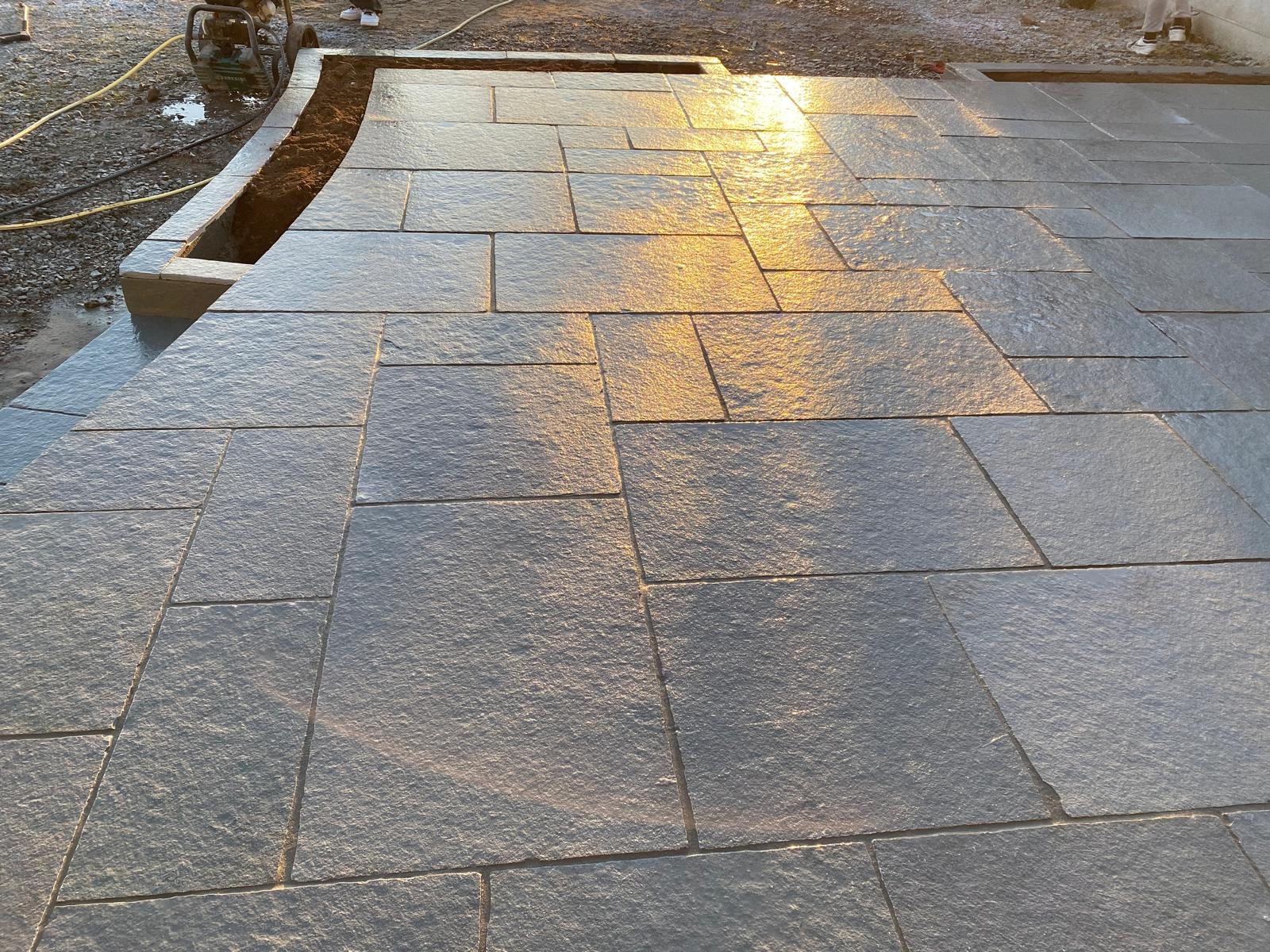 Freshly laid grey sandstone patio in random sizes catching low golden sunlight, with a curved edge course framing a new planting bed