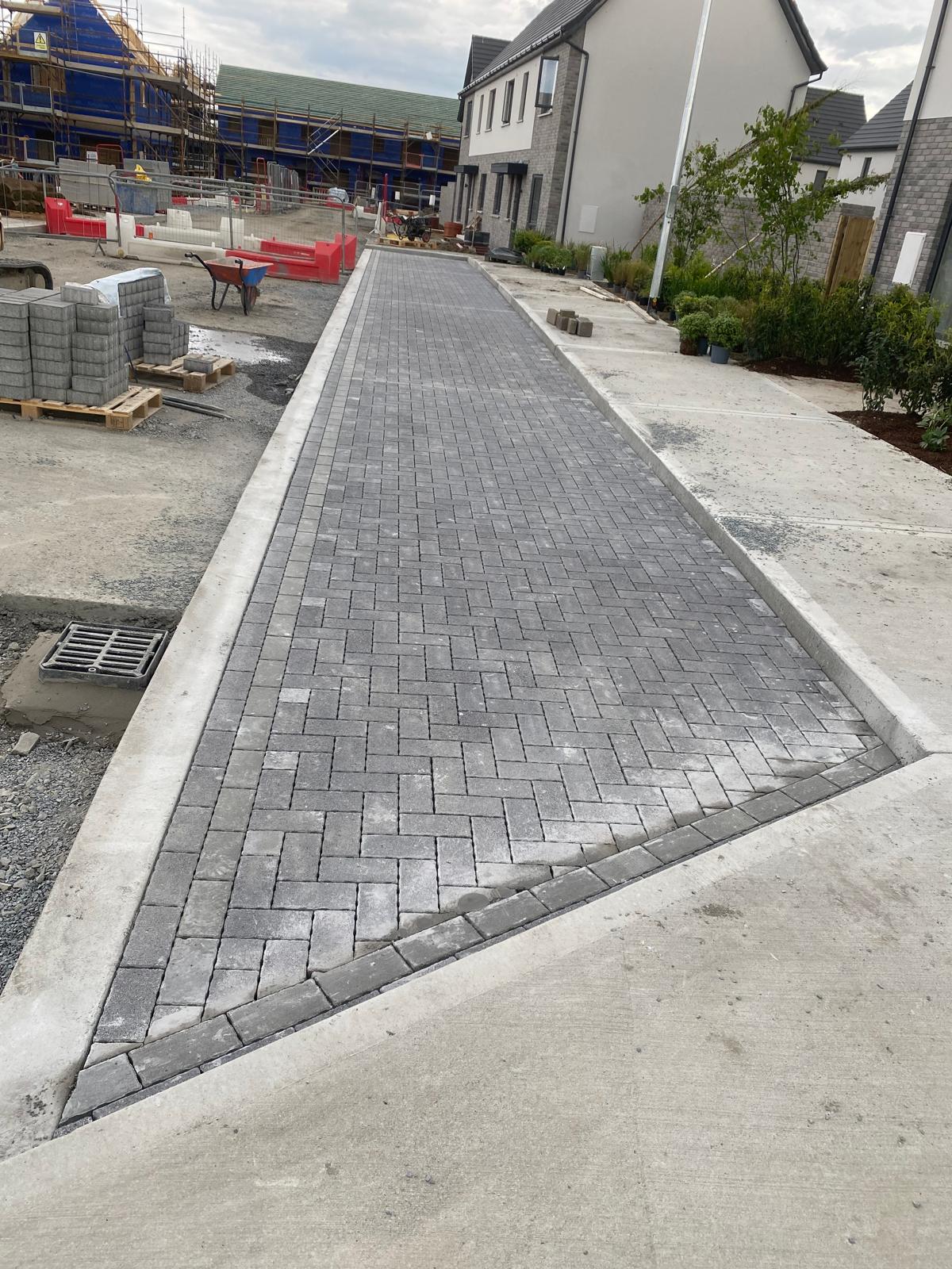 Long charcoal herringbone block paved path with a darker soldier course edge and concrete kerbs running between houses on a construction site