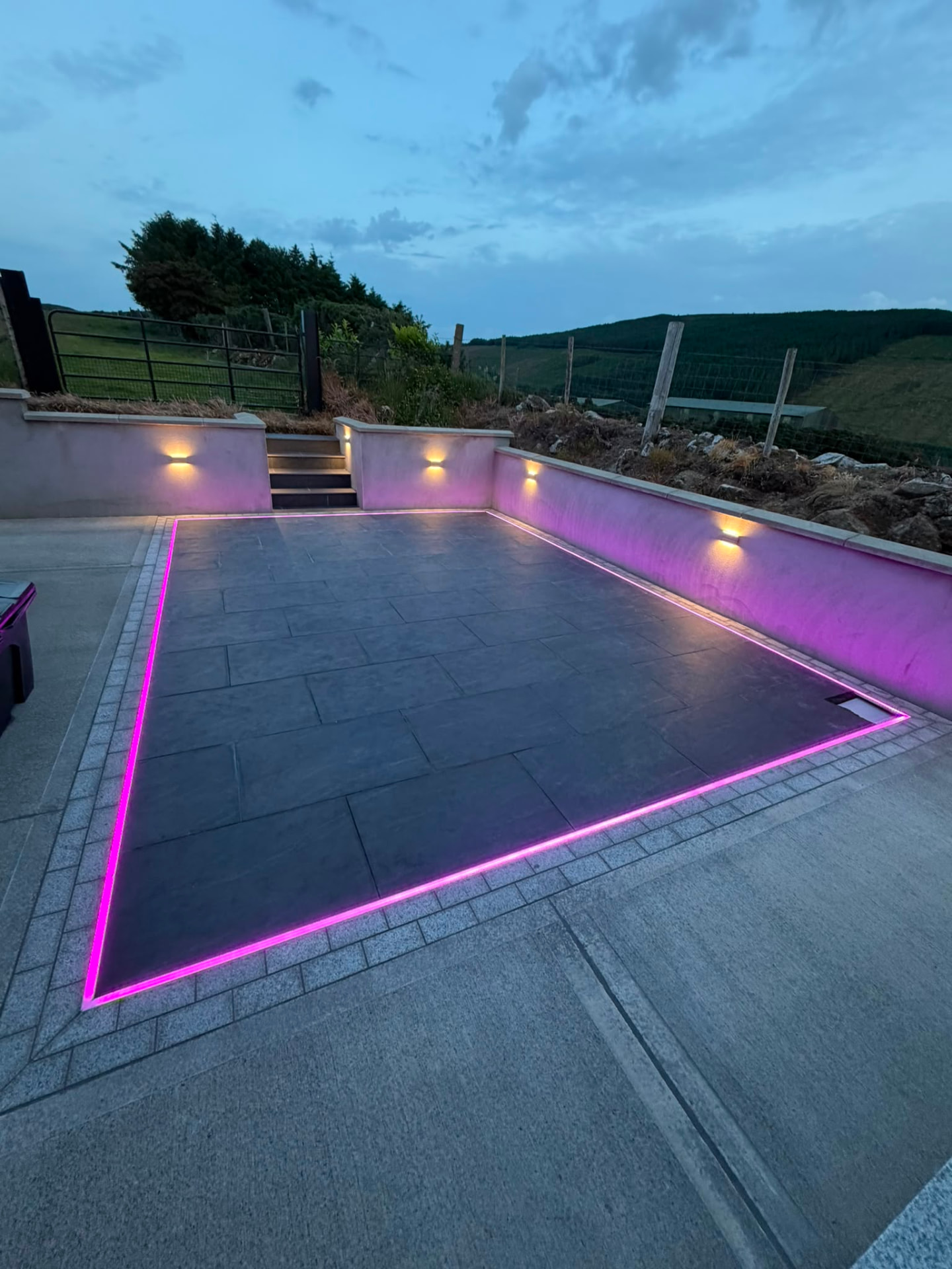 The same dark paved patio at blue hour with the integrated perimeter light line set to a pink colour accent and warm wall lights along the retaining walls