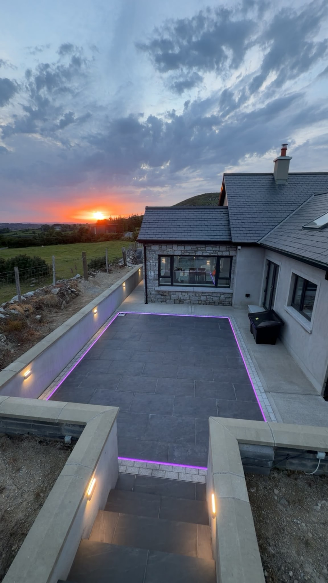 Elevated view of a sunken paved patio at sunset with an integrated light line around the paving, lit steps and wall lights, beside a stone and rendered house