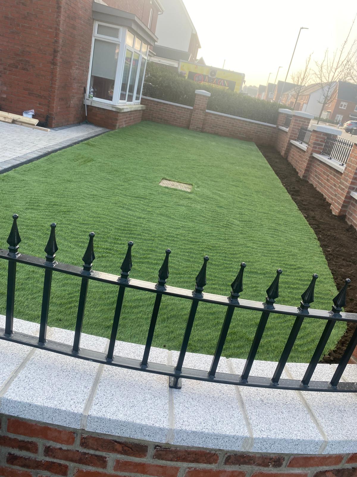 Freshly laid front lawn enclosed by a brick boundary wall with light granite capping and black railings, beside block paving at the house