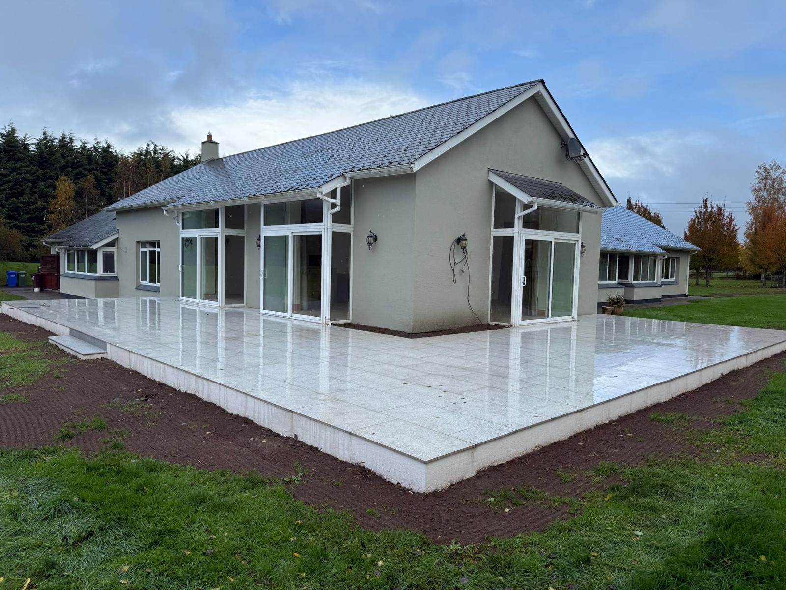 Large light granite slab patio wrapping around two sides of a bungalow, wet from rain, with a capped edge course and a low step at one corner