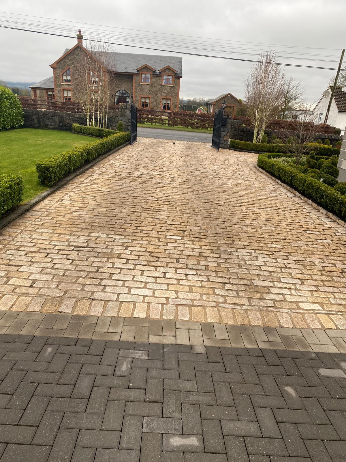 Golden granite cobble driveway running from open black gates towards a two storey house, framed by clipped box hedging and lawn