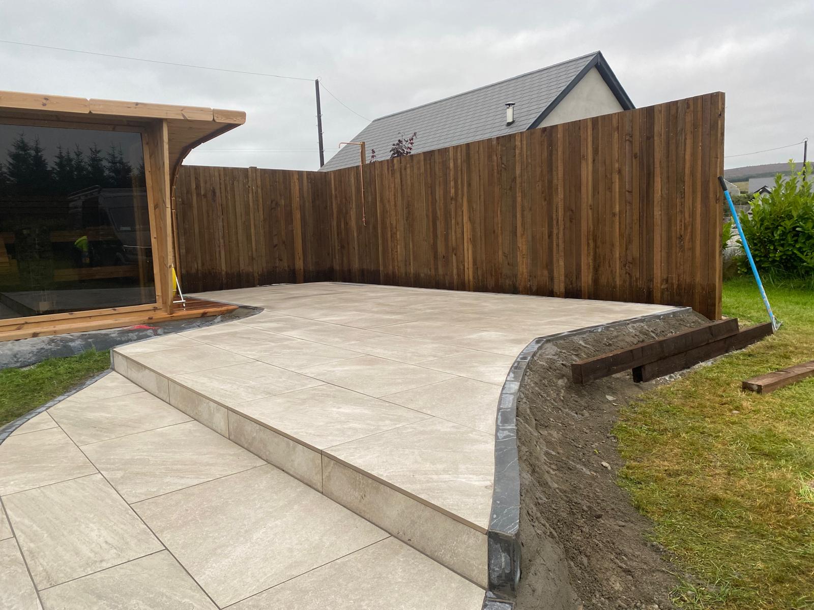 Raised pale porcelain patio with a sweeping curved edge, dark kerb detail and a single wide step down, beside a timber garden room and stained fencing