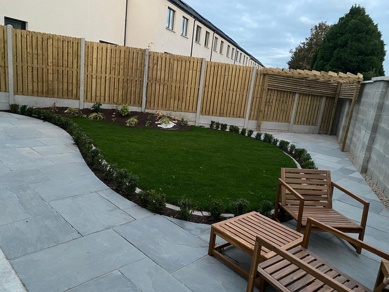 Grey sandstone patio curving around a shaped lawn with clipped low hedging, planted borders, timber fencing and a slatted pergola