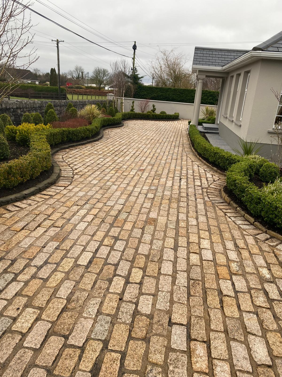 Sweeping granite cobble driveway with curved kerb edging, clipped hedging and mixed planting leading to a house with a pillared porch