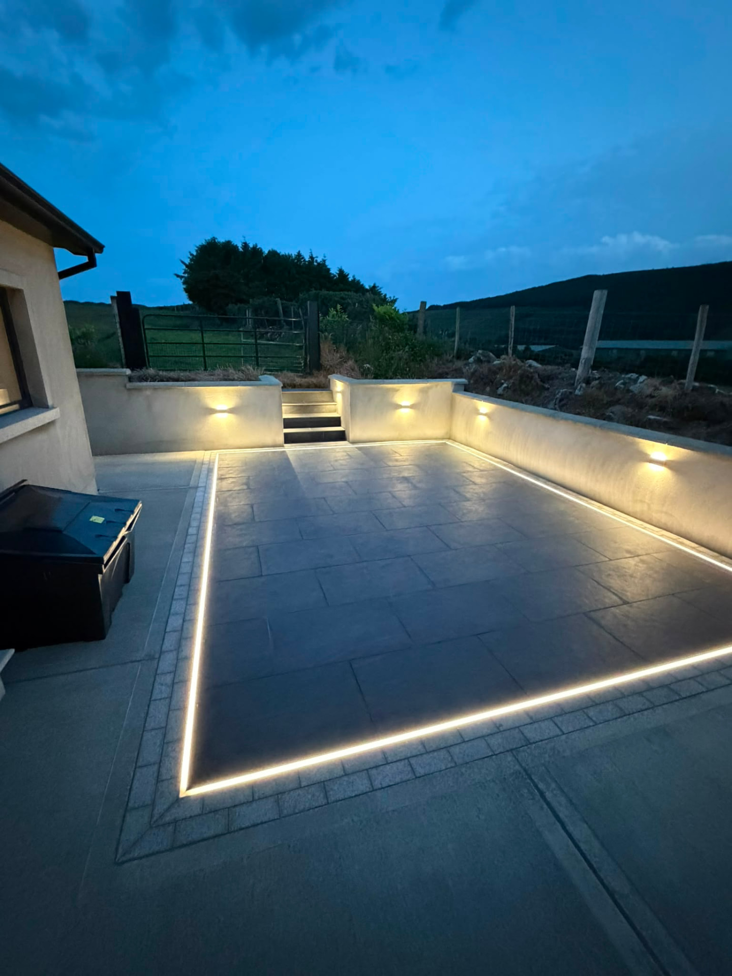 Dark large format paved patio at dusk with a continuous warm white light line set into the perimeter joint and wall lights washing the rendered retaining walls