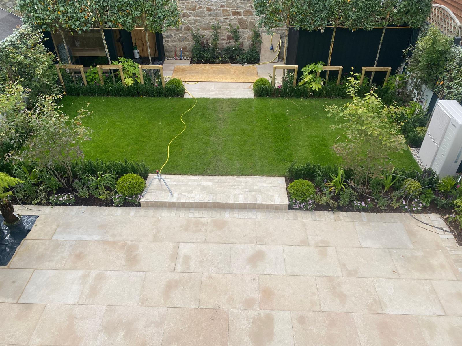 Elevated view of an enclosed courtyard garden with a pale limestone patio, a low paved step, a level lawn and clipped planting borders along both sides