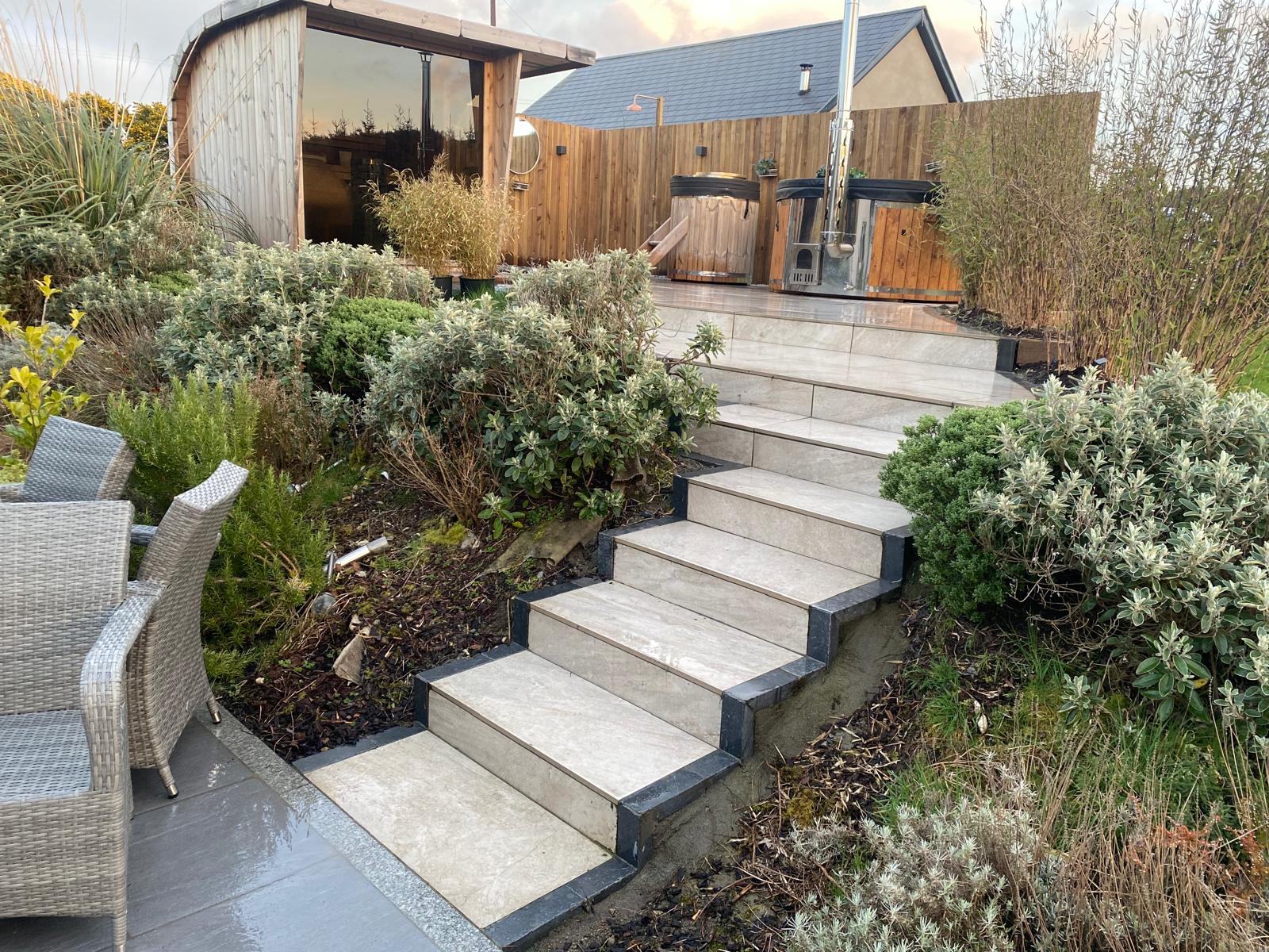 Flight of pale porcelain steps with dark contrasting nosings rising between established shrub planting to an upper paved terrace with a timber garden room