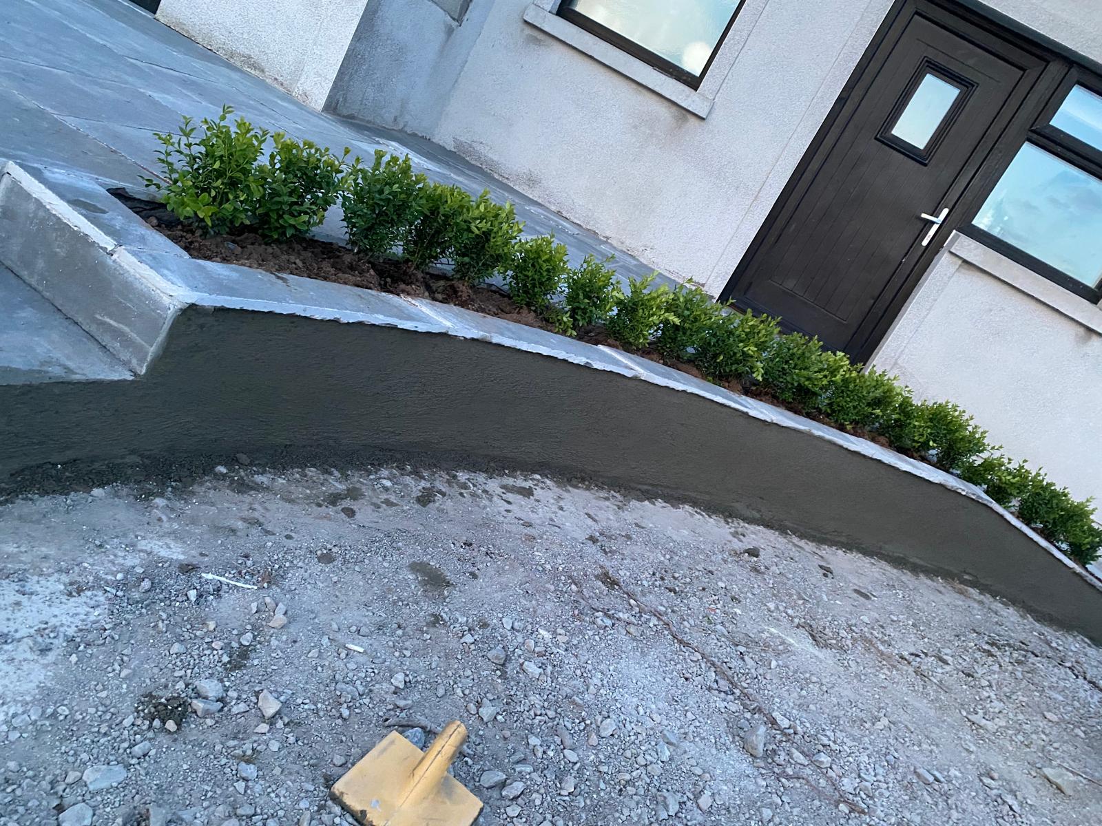Newly rendered curved retaining wall with a capped top holding a narrow planting bed of clipped shrubs alongside paving at a house entrance