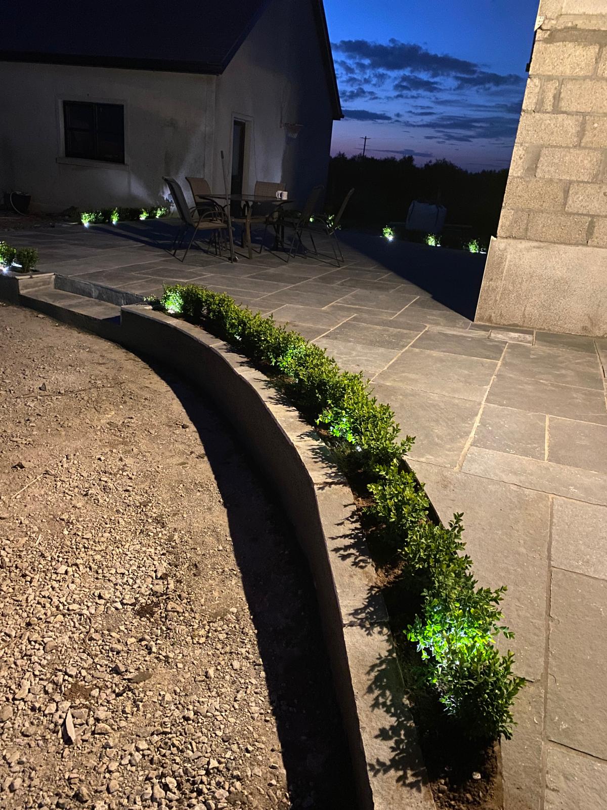Evening view of a sandstone patio with a curved rendered retaining wall, a row of clipped shrubs and small spotlights lighting the planting