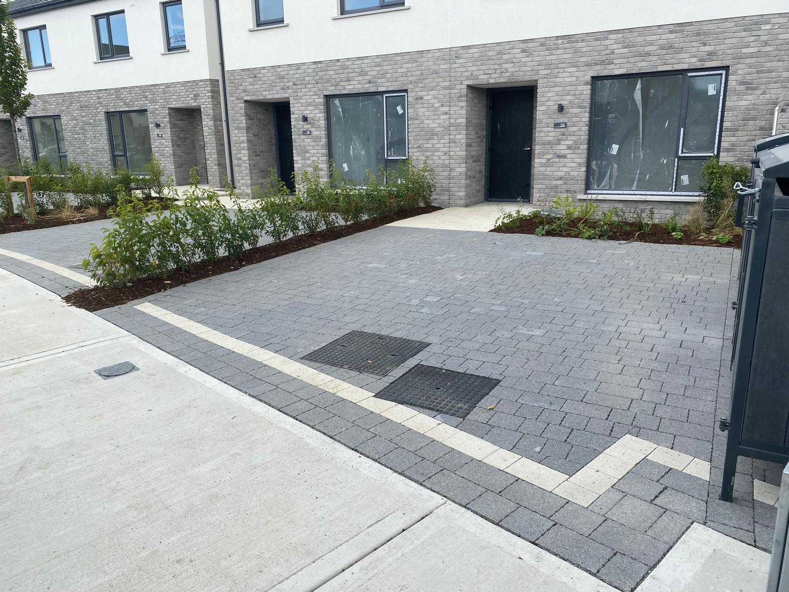 Grey block paved driveway and parking area with a light border course turning at the entrance, serving a row of new brick and rendered houses with fresh planting beds