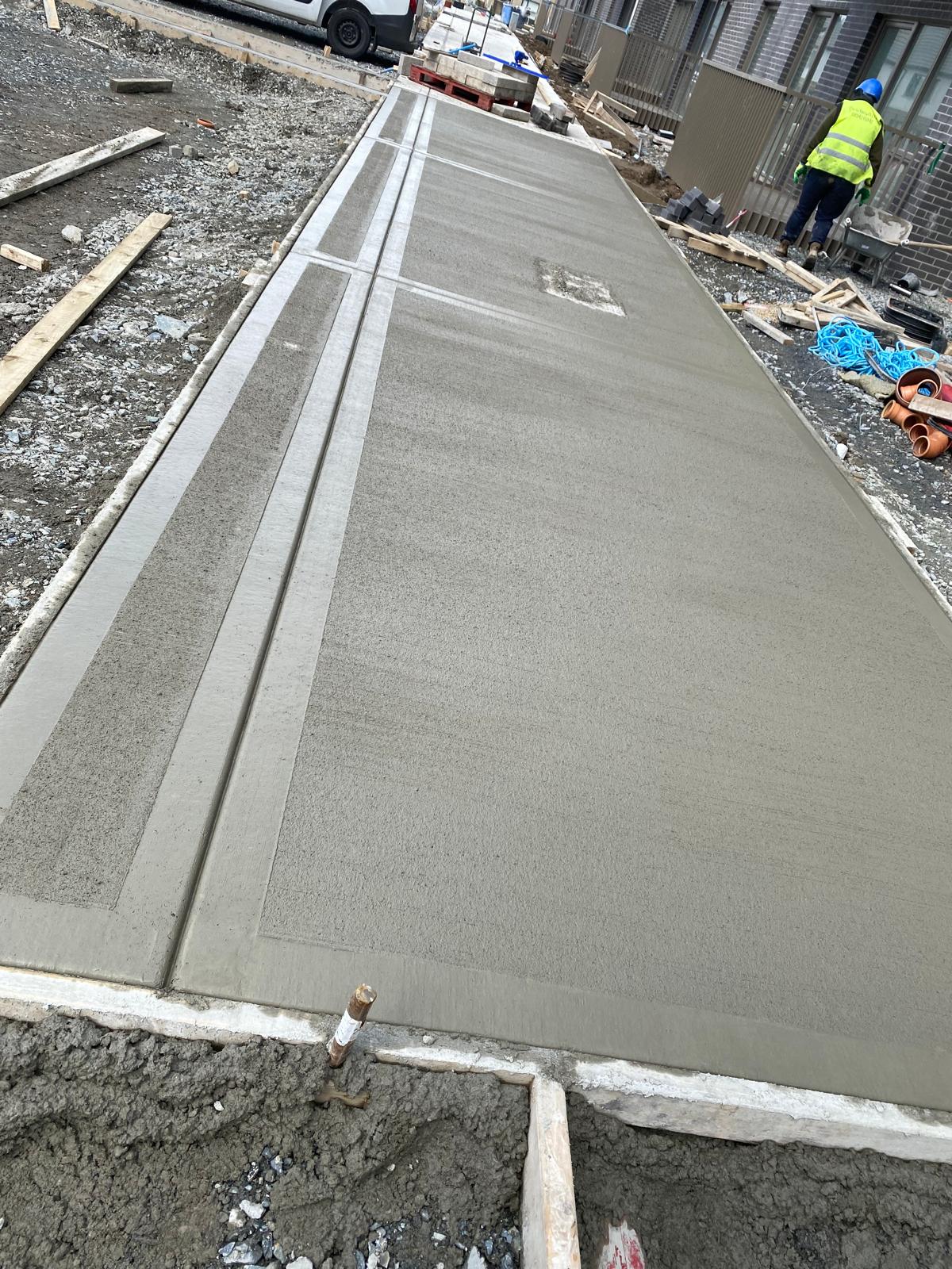 Freshly poured concrete path with a brushed finish and formed edge channels running alongside brick houses on a construction site