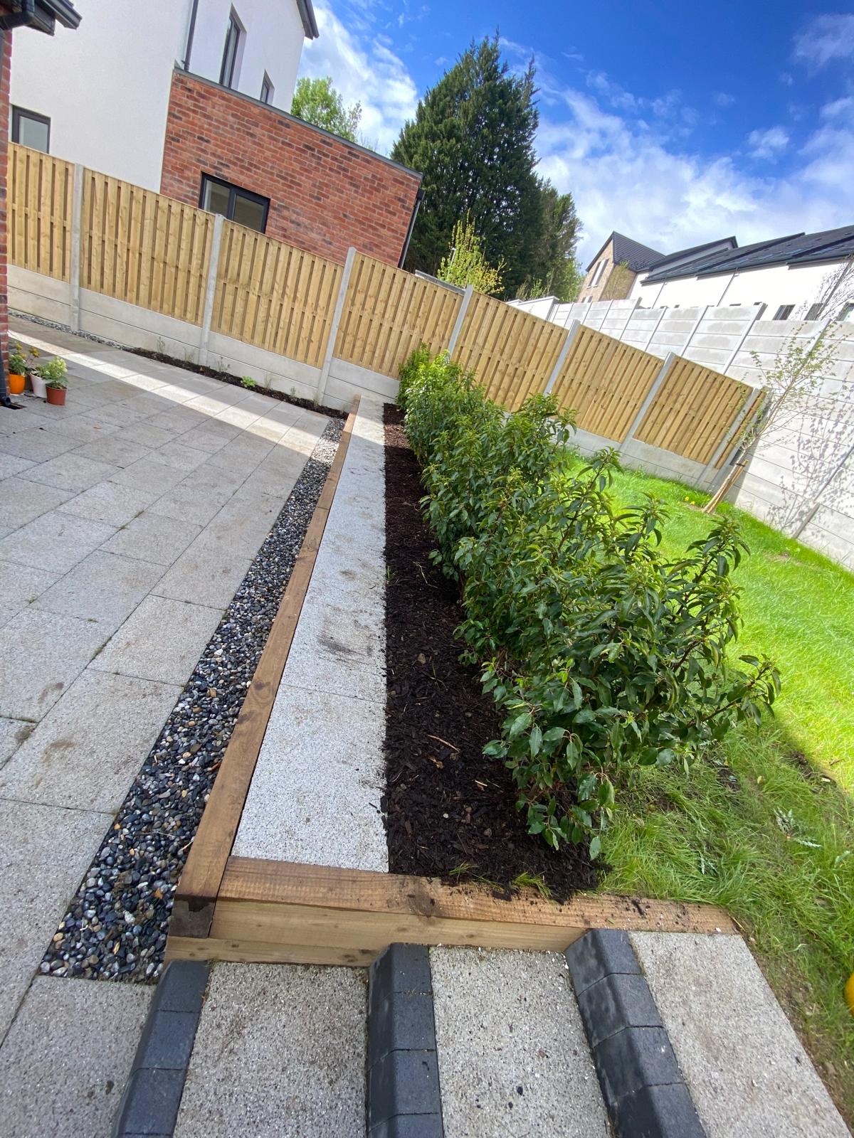 Timber sleeper edged planting bed of young shrubs beside a granite paved path, a pebble drainage channel and lawn, enclosed by timber fencing