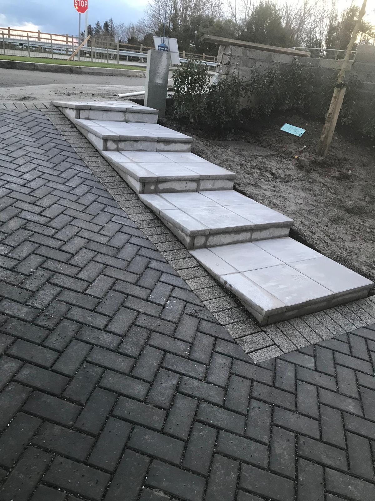 Flight of wide paved steps with slab treads and block risers rising alongside charcoal herringbone block paving at a site entrance