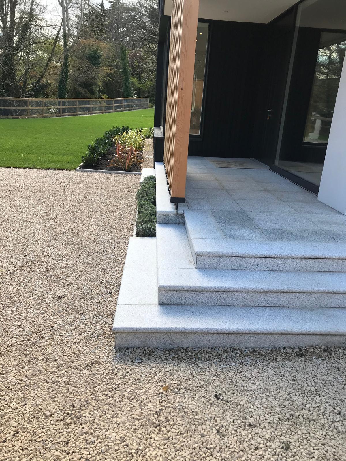 Wide granite entrance steps and landing beside a gravel area, with low clipped planting and a dark timber clad entrance