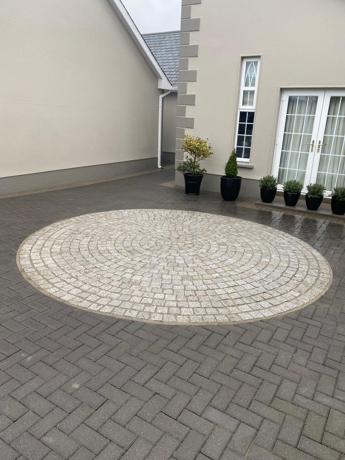 Circular pale granite cobble feature set into a charcoal herringbone block paved courtyard beside a rendered house with potted shrubs
